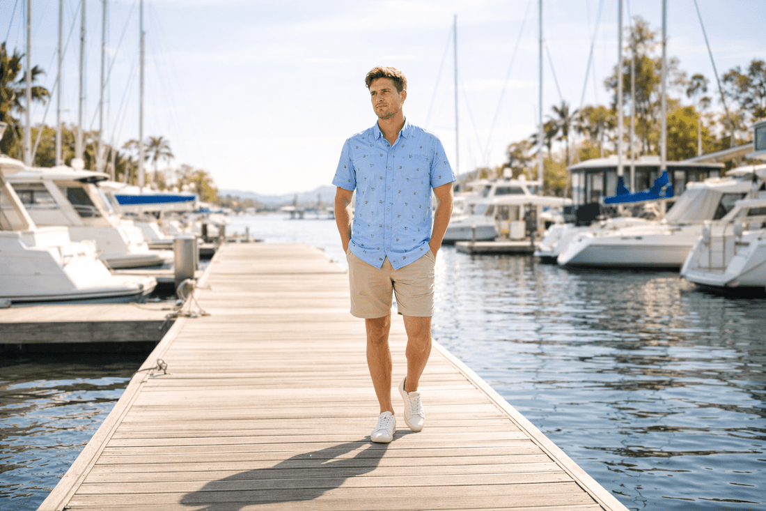 Full-body lifestyle shot of a man walking on a marina dock in bright morning sunlight, wearing a light blue short-sleeve printed shirt and beige shorts, with boats and calm water in the background.