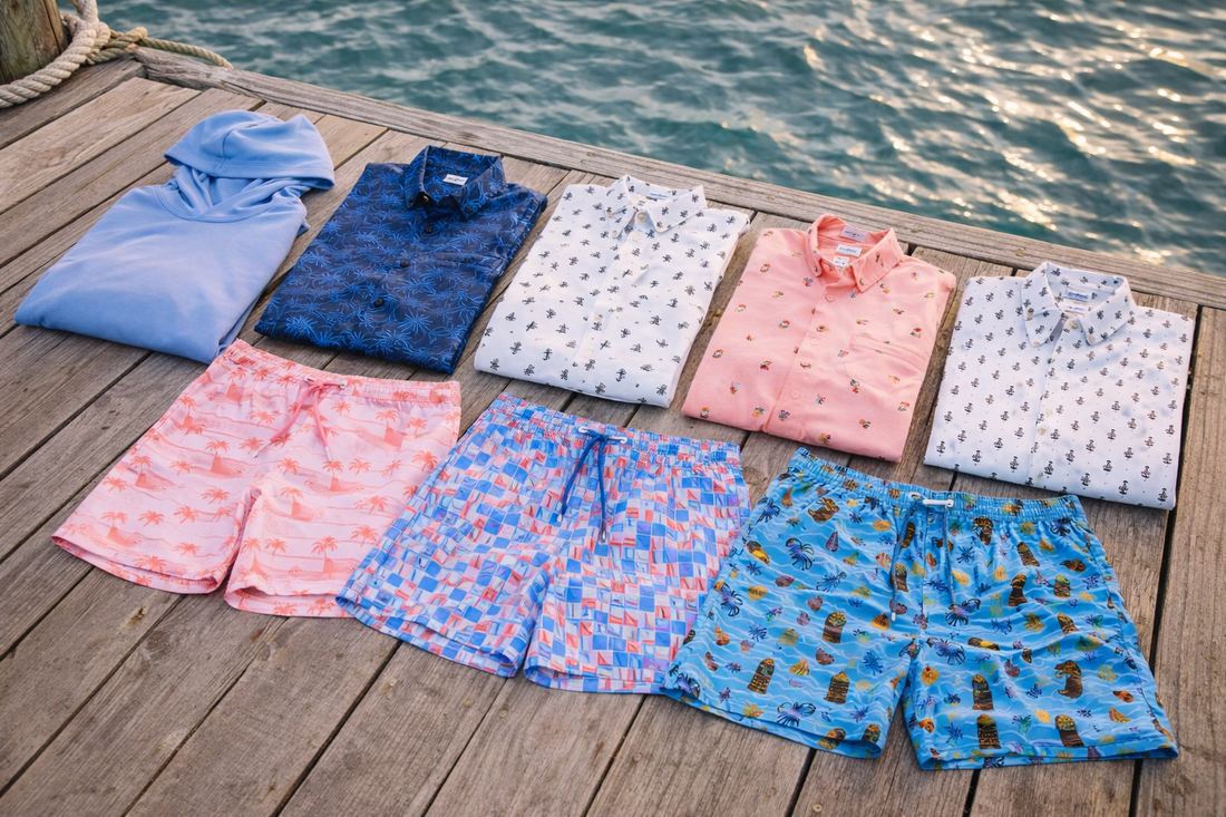 Men’s spring coastal apparel collection arranged on a weathered marina dock, featuring printed short-sleeve woven shirts, colorful swim trunks, and lightweight layering pieces beside sunlit rippling water.