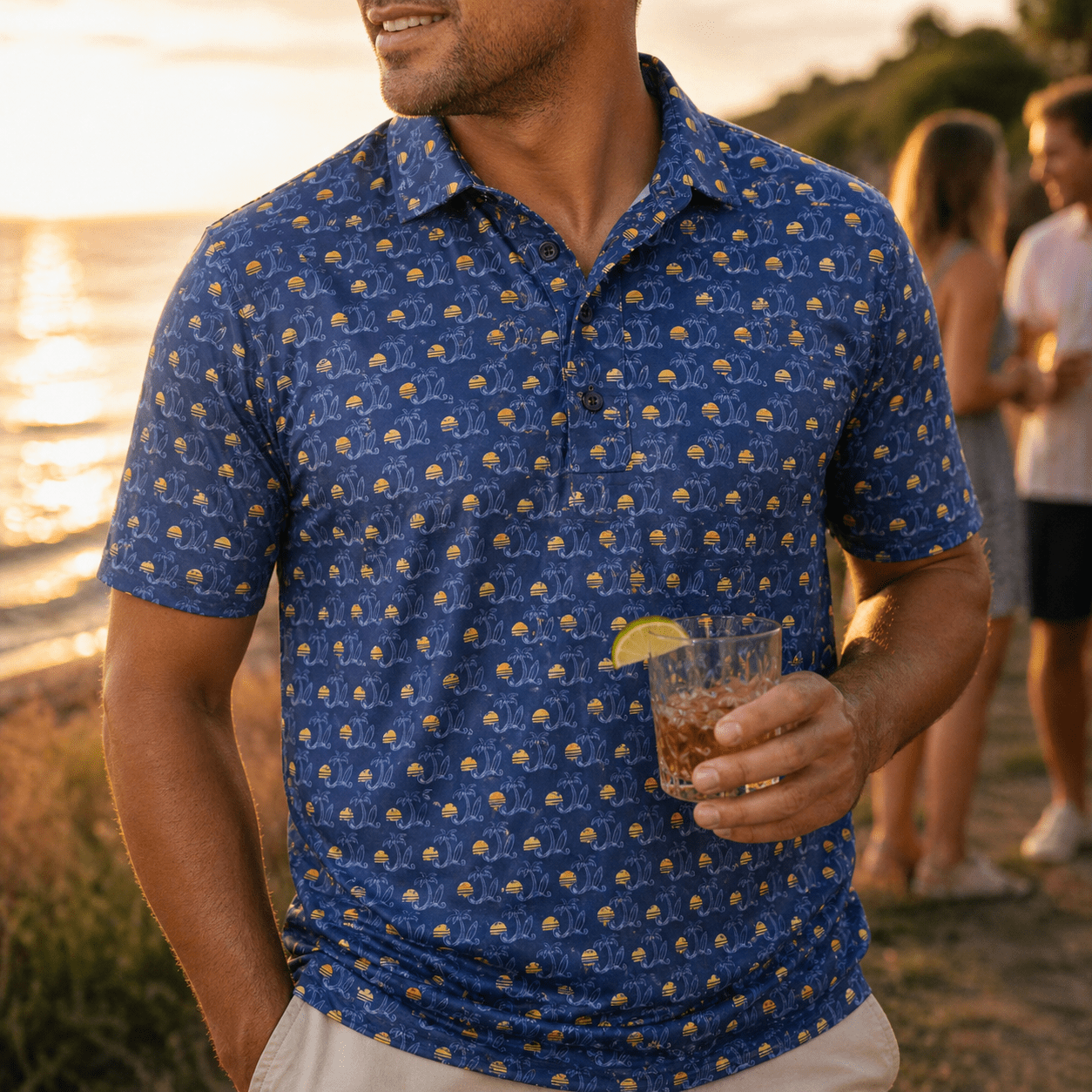 Man wearing a blue patterned shirt holding a drink by a lake with people in the background