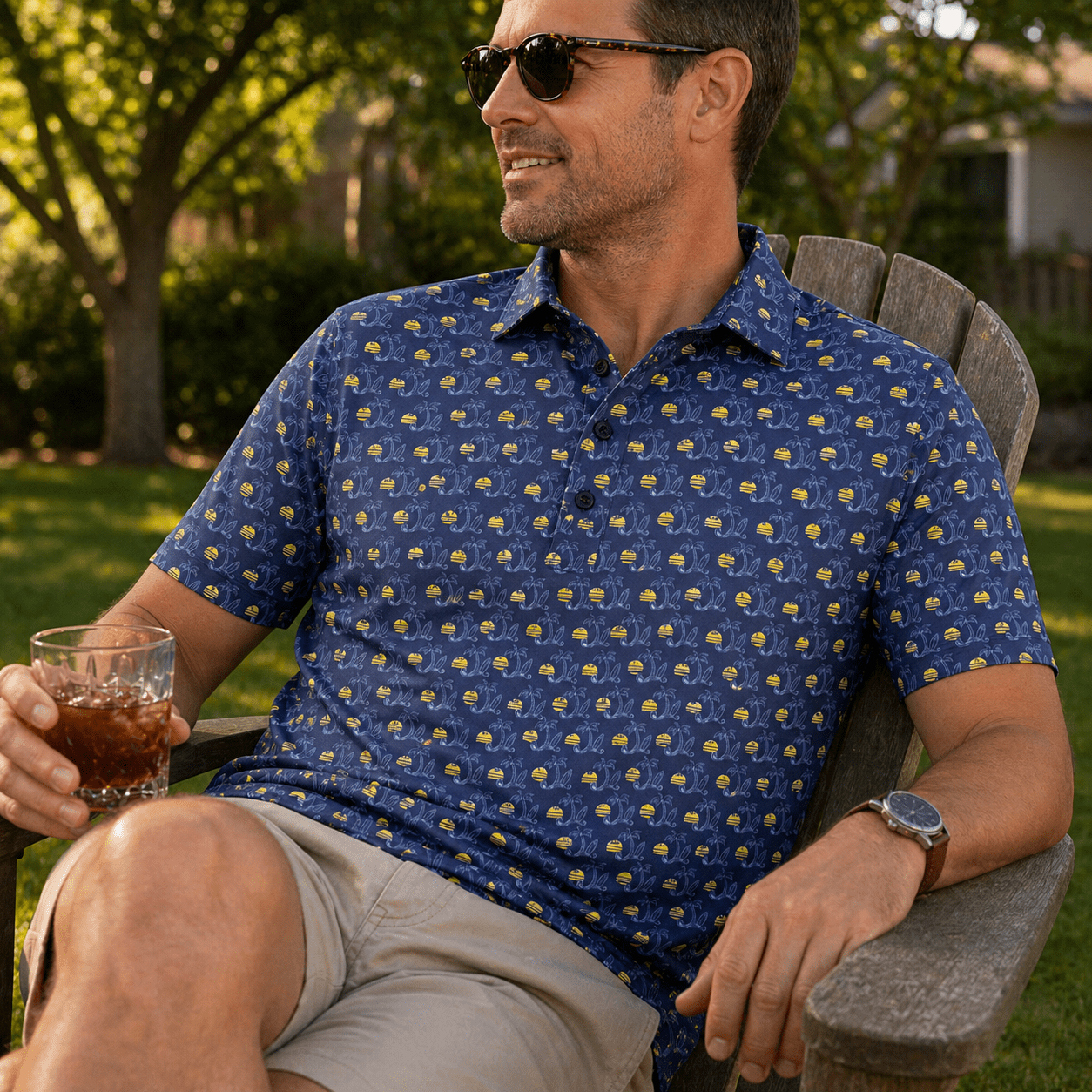 Man sitting outdoors in a garden, wearing a blue patterned shirt and sunglasses, holding a drink.